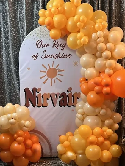 A cute "Our Ray of Sunshine" themed birthday decoration. This setup features a personalized arched backdrop and a warm-toned balloon garland in shades of orange and yellow.