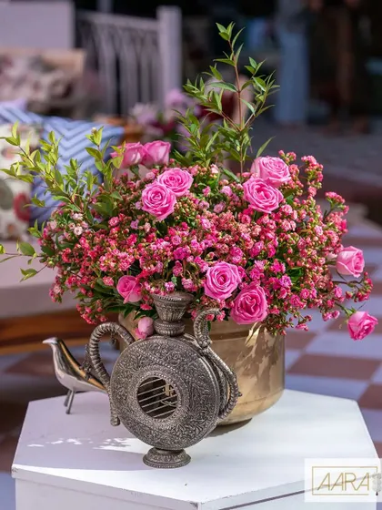 Details from our 'Pomegranate Spring' theme, where wild flowers and bright colors meet quirky artifacts. A vintage silver flask sits beside a brass vase overflowing with pink roses, creating a charming vignette.