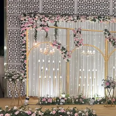 An elegant stage backdrop featuring golden arches, pink floral arrangements, and candle-like lights against a white curtain. This design is perfect for a romantic engagement or sangeet.