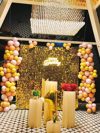 A glamorous first birthday celebration featuring a brilliant gold sequin wall. The backdrop is flanked by organic balloon garlands in peach and yellow, with elegant pedestals and a large number one foil balloon completing the look.