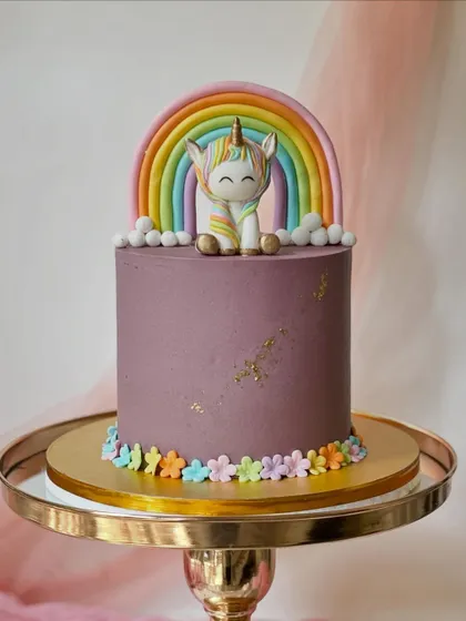 A modern unicorn cake with a rich purple buttercream. The design is completed with a fondant rainbow, a cute unicorn figure, and a border of tiny flowers.