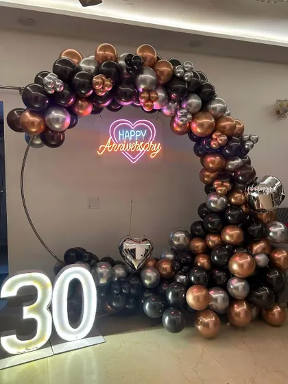 Milestone anniversaries deserve a special celebration. This setup for a 30th anniversary features a stylish circular balloon arch in black, silver, and rose gold, a 'Happy Anniversary' neon sign, and large illuminated marquee numbers.