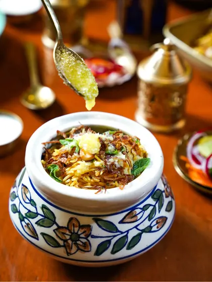 A rich and aromatic biryani in a traditional handi, with a spoonful of ghee being drizzled on top. The action and the warm lighting create a mouth-watering image for an Eid celebration.