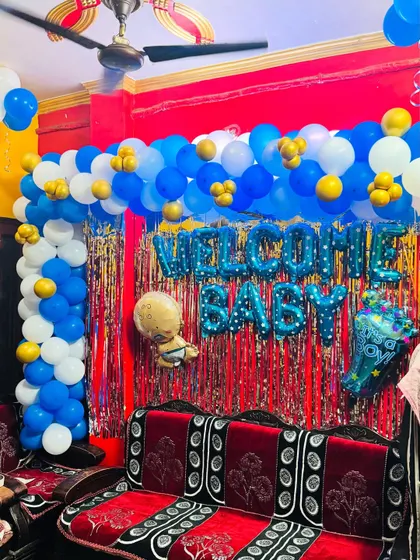 A 'Welcome Baby' decoration in a living room. The setup includes a blue, white, and gold balloon arch over a red shimmer curtain.