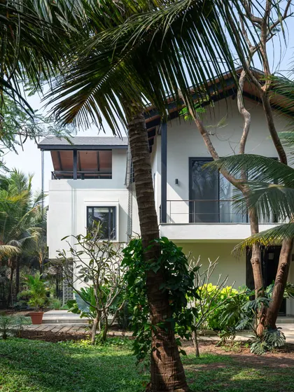 An exterior view of Grove House, a weekend retreat in Alibaug surrounded by a lush coconut plantation. The design uses a simple palette of white walls and dark trim, allowing the vibrant tropical foliage to take center stage.