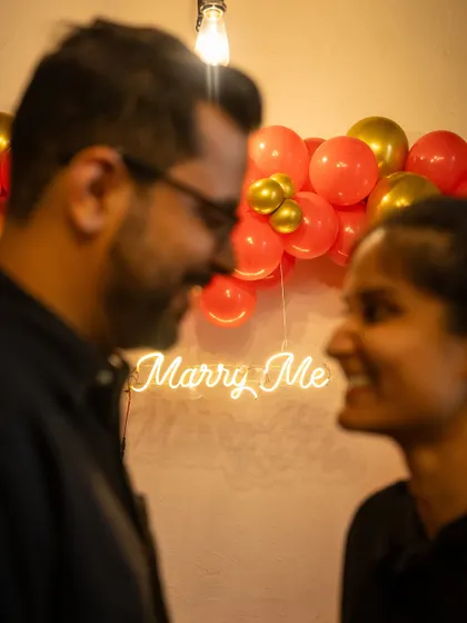 A close-up shot capturing the intimacy of a proposal. The soft glow of the 'Marry Me' neon sign creates a romantic and personal moment between the couple.