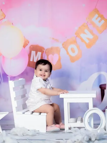 This baby girl is celebrating her first birthday with a "Mr & Mrs ONE" banner in the background, sitting at a tiny white table.