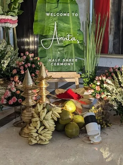 A welcome setup for a half-saree ceremony featuring a custom sign on a banana leaf, surrounded by traditional elements like brass lamps, coconuts, and fresh flowers.