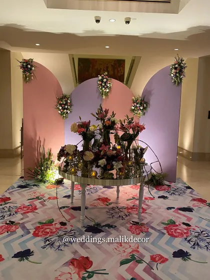 A creative floral installation for a Mehendi event. A mirrored table serves as the base for a stunning arrangement, set against a custom-built backdrop of curved pastel panels for a modern, artistic look.