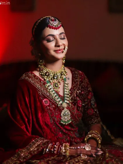 A serene portrait of the bride with her eyes closed, taking a moment for herself. The warm, dramatic lighting beautifully illuminates her red velvet lehenga and heavy bridal jewelry.