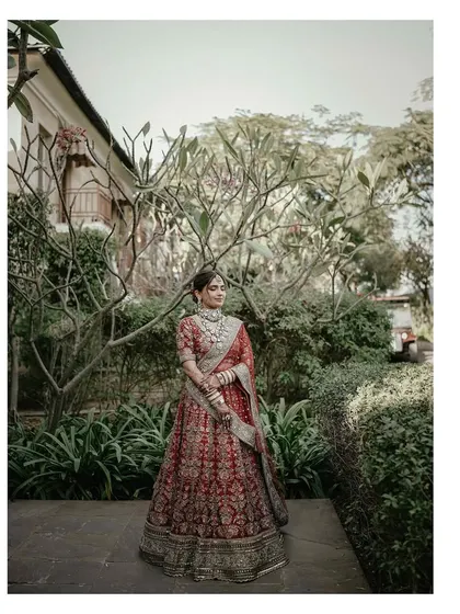 A full-length portrait of the bride in her red lehenga, standing in a garden, the picture of classic bridal beauty.