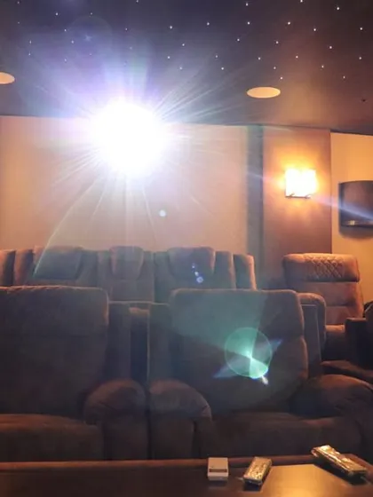 The projector beam creates a starburst effect, illuminating the room and highlighting the warm, cozy atmosphere of this well-appointed home theater.