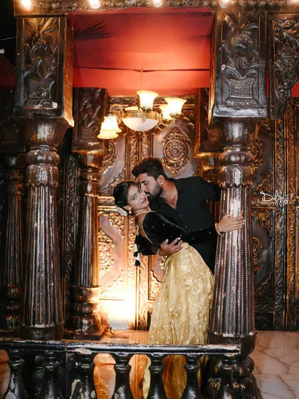 A romantic and glamorous shot on a balcony-like set, with the couple framed by ornate pillars and warm lighting.
