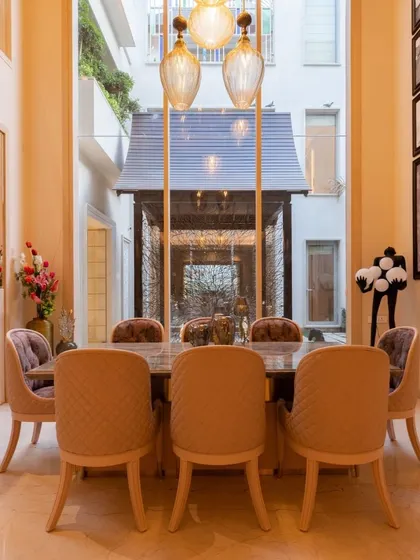 A double-height dining area creates a sense of grandeur and openness. The space is elegantly appointed with quilted chairs and pendant lighting, with a view into a serene courtyard that enhances the dining experience.
