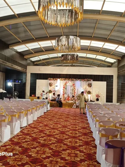 This wide shot shows a complete hall setup for a wedding ceremony. You can see the main stage decor, the aisle carpet, and the neat arrangement of chairs with covers, all ready for the guests to arrive.