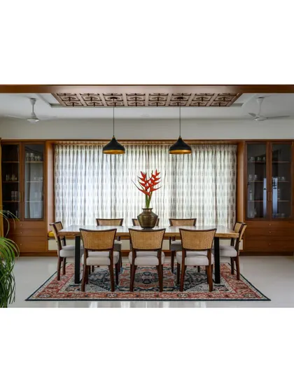 The 'Vasudhalayah' dining area features a live-edge table anchored by an intricately carved wooden ceiling panel. The space is flanked by two crockery units and features a bay window seating nook.