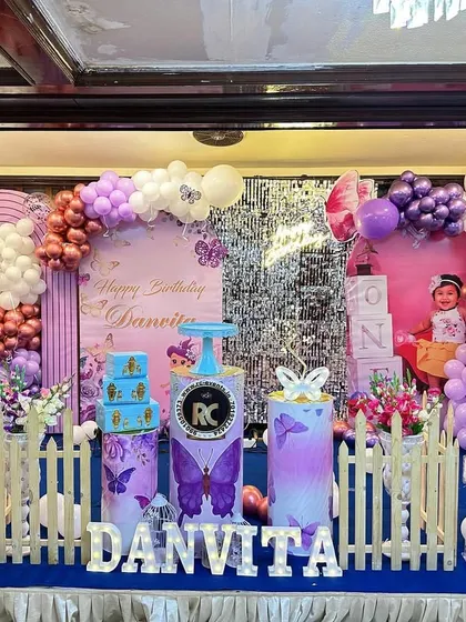 A full view of a butterfly-themed stage decoration with personalized photo cutouts and a purple and metallic balloon arrangement. The setup includes themed plinths and light-up letters for the name.