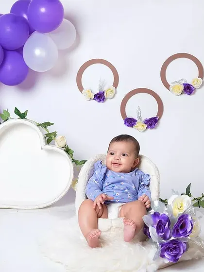 A sweet baby girl sitting in a heart-shaped bowl, surrounded by a cascade of purple and white balloons.