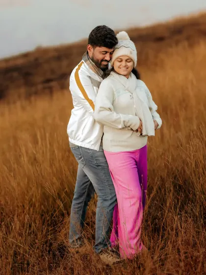 An expecting couple walks hand in hand through a rustic field, their comfortable winter outfits adding to the cozy, natural feel of the moment. I encourage simple, candid actions like walking and talking to capture authentic connection.