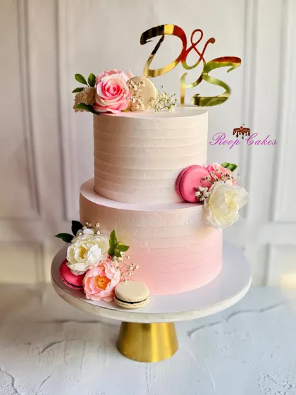 Layers of love in blush, blooms, and gold. This two-tier engagement cake features a soft pink ombre, fresh flowers, and delicate macarons, personalized with the couple's initials on a gold topper.
