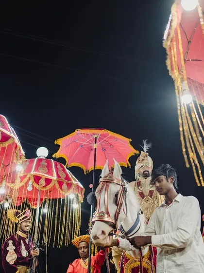 A royal baraat procession, with the groom on a white horse, surrounded by an entourage carrying ornate, lit-up umbrellas.