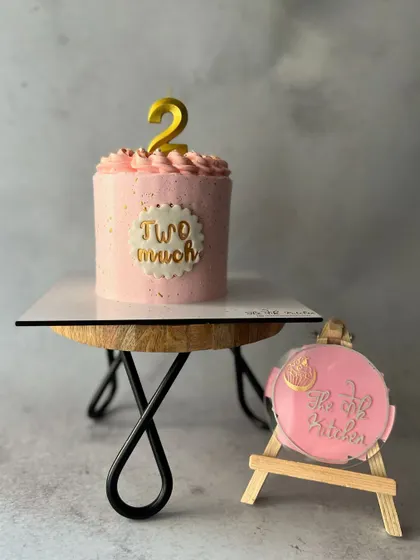 A simple and chic mini cake for a second birthday, with the playful message "Two Much" written on a fondant plaque.