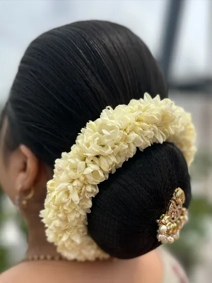 A classic South Indian bun or `kondai`, neatly wrapped and adorned with a circle of fresh jasmine flowers and a simple jewel. This is a fundamental style for any bridal artist.
