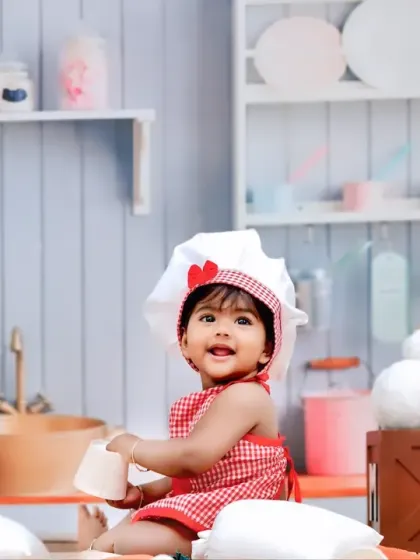 Our little chef sits happily in a fully equipped miniature kitchen set, complete with pastel-colored utensils, a tiny sink, and shelves.