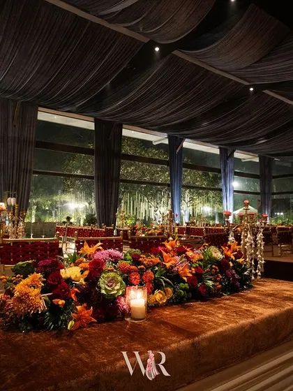 A long dining table covered in rich brown velvet, adorned with a vibrant floral runner of red, orange, and green. The setting is opulent and colorful, set under a ceiling of dark blue drapes and chandeliers.