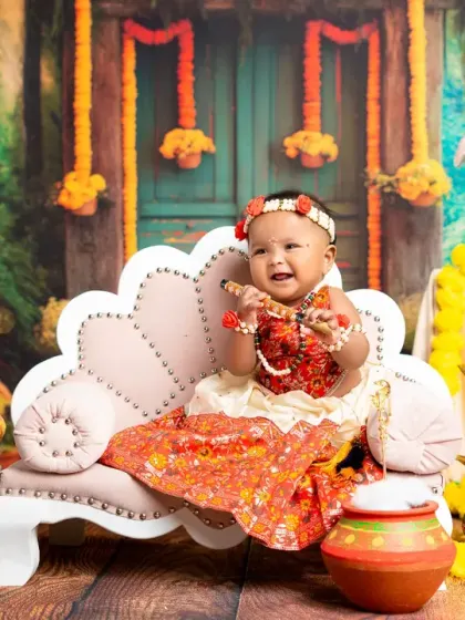A joyful baby girl dressed in a beautiful traditional outfit for a Janmashtami-themed photoshoot. The set is decorated with marigolds, a peacock, and clay pots to create an authentic atmosphere.