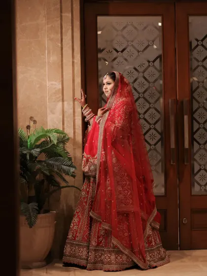 A moment frozen in time, a heart filled with love. The bride stands by a doorway, her red lehenga a vibrant splash of color.