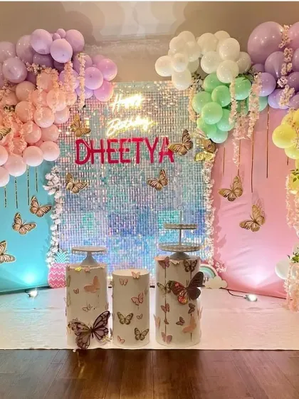 A vibrant butterfly-themed party featuring a stunning iridescent shimmer wall that catches the light beautifully. The setup is framed by a pastel rainbow balloon garland and includes themed plinths for the cake.
