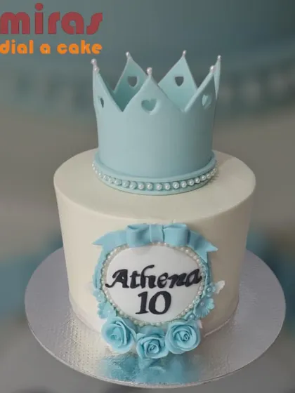 A regal cake for a 10th birthday, topped with a light blue crown. The design is accented with blue roses and a personalized name plaque, fit for a young queen.