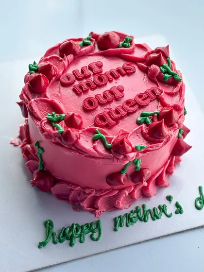 A special cake for Mother's Day with the message "Our Moms Our Queen." The vibrant pink buttercream is decorated with piped cherries for a fun and festive look.