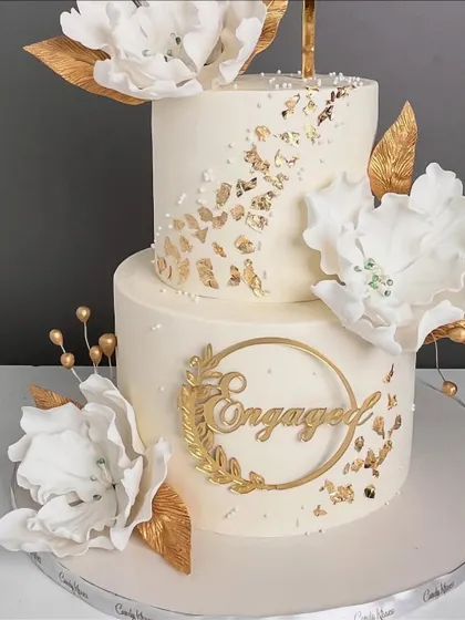 A beautiful two-tiered engagement cake in white and gold, featuring large sugar flowers, gold leaf, and an elegant "Engaged" topper.