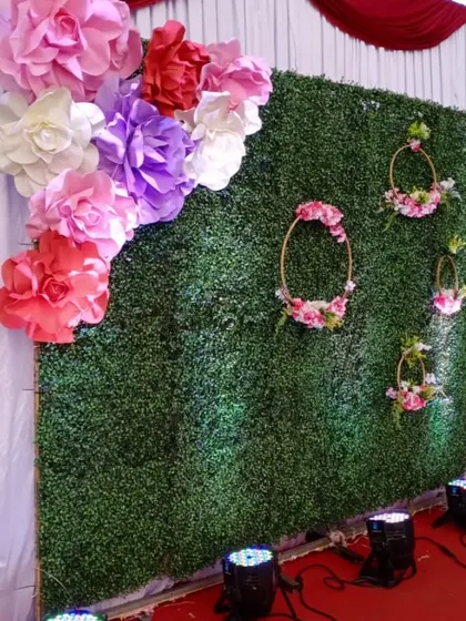 A fresh and vibrant photo wall for a ring ceremony. This backdrop uses an artificial green grass wall as a base, decorated with large, colorful paper flowers and delicate floral hoops for a garden-party feel.