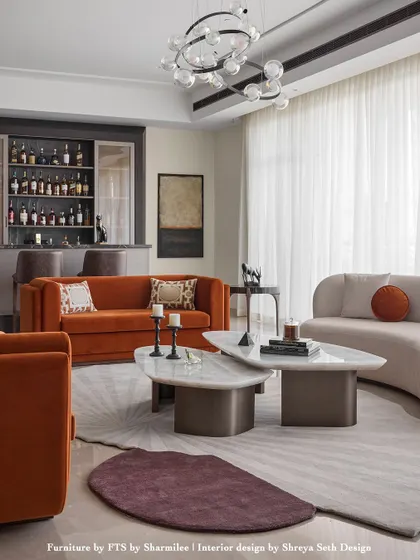 A vibrant living room where color and form create a lively dialogue. The burnt orange sofa provides a bold focal point, complemented by a curved neutral sofa and uniquely shaped marble coffee tables. A custom bar is seamlessly integrated into the background, making this a perfect space for entertaining.