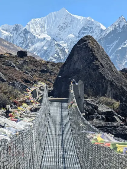 A suspension bridge adorned with colorful prayer flags leads towards a massive, snow-covered peak in the Langtang region of Nepal.