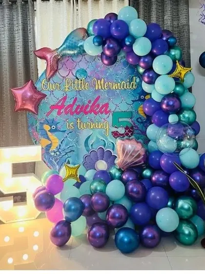 A close-up of the "Our Little Mermaid" 5th birthday setup. The decoration features a custom backdrop, a marquee number '5', and a balloon arch with star and shell accents.