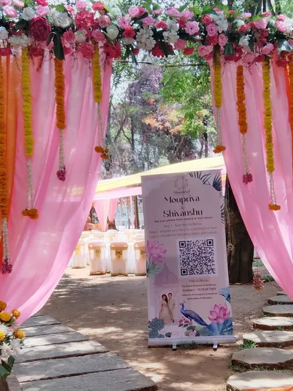 A welcoming entrance to an outdoor ceremony, with pink drapes, floral garlands, and a custom welcome sign with a QR code.