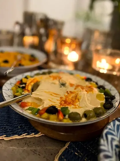 A cozy, candlelit shot of our creamy hummus topped with olives and peppers. This is a staple of our festive and Mediterranean spreads, perfect for any unforgettable dinner.