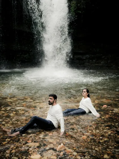 A wide shot of the couple sitting peacefully in the water at the base of a waterfall. This image has a serene and almost meditative quality, capturing a moment of shared tranquility.