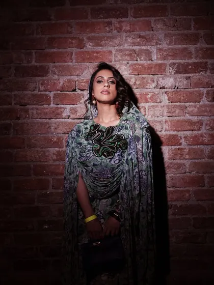 A full-length view of the Anamika Khanna x H&M look. The hair is styled with soft curls, and the makeup is bold enough to stand out against the patterned fabric and dramatic silhouette of the cape.