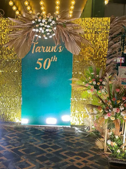 A stylish 50th birthday photo booth combining a gold shimmer wall with a teal panel and beautiful pampas grass floral arrangements.