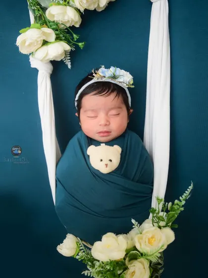 A creative newborn portrait where the baby is swaddled and appears to be resting in a soft white sling, decorated with white flowers against a deep teal background.