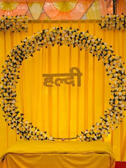 A classic Haldi stage setup with a circular floral arch against a bright yellow backdrop. The stage is covered with a red carpet for a pop of color.
