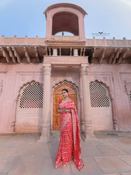 A wide-angle shot showcasing a woman in a stunning red saree against a grand pink heritage building. This photo captures the majestic scale of the location and the elegance of the attire.