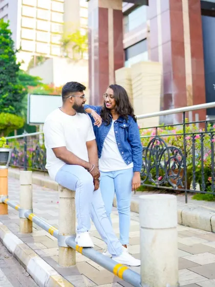 A casual pre-wedding photo in a modern urban setting. The couple is sitting on a guardrail, creating a relaxed and contemporary vibe.