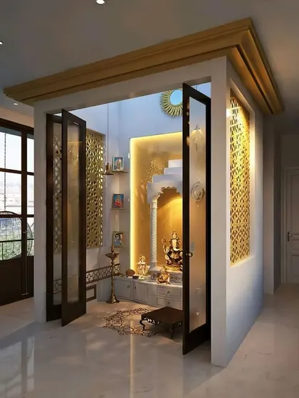This pooja room is designed as a separate enclosure with foldable glass doors. The golden accents and intricate jaali work on the back wall create a rich and sacred feel.