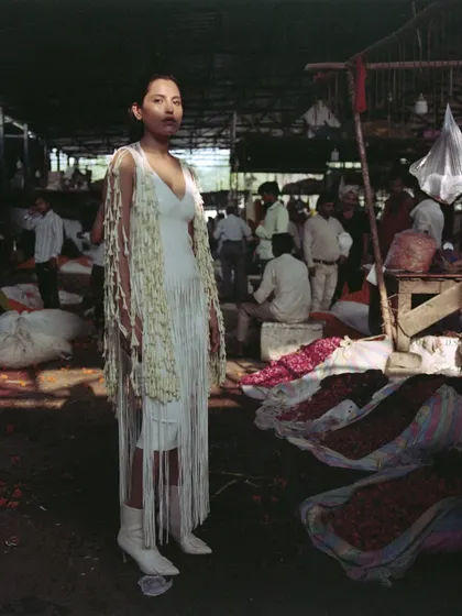 A full-length look from the 'Phool Mandi' editorial. The styling features a white fringed dress layered with a jacket made of jasmine flowers, blending fashion design with natural elements in a raw, market environment.